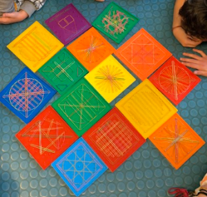 Studentesse e studenti formano figure geometriche utilizzando fili tesi attorno a teste di chiodi fissati su tavolette colorate.