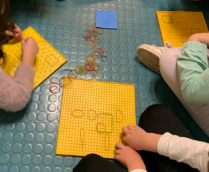Studentesse e studenti formano figure geometriche utilizzando fili tesi attorno a teste di chiodi fissati su tavolette colorate.