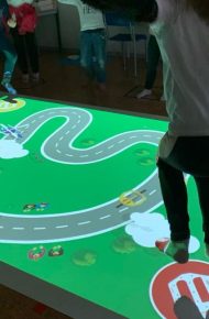 Su una superficie rettangolare di circa 8 metri quadrati su cui viene proiettato un tabellone di gioco, una bambina e un bambino toccano dei pulsanti con i piedi per controllare dei veicoli su un circuito