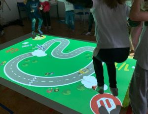 Su una superficie rettangolare di circa 8 metri quadrati su cui viene proiettato un tabellone di gioco, una bambina e un bambino toccano dei pulsanti con i piedi per controllare dei veicoli su un circuito