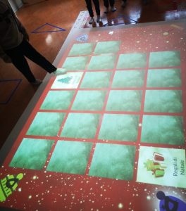 Su una superficie rettangolare di circa 8 metri quadrati su cui viene proiettato un tabellone di gioco, bambine e bambini uno a turno toccano con i piedi tessere di memory a tema natalizio.