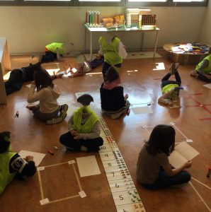 Bambine e bambini misurano il perimetro di forme geometriche, formate con nastro adesivo sul pavimento, utilizzando la colla e il proprio pollice come unità di misura